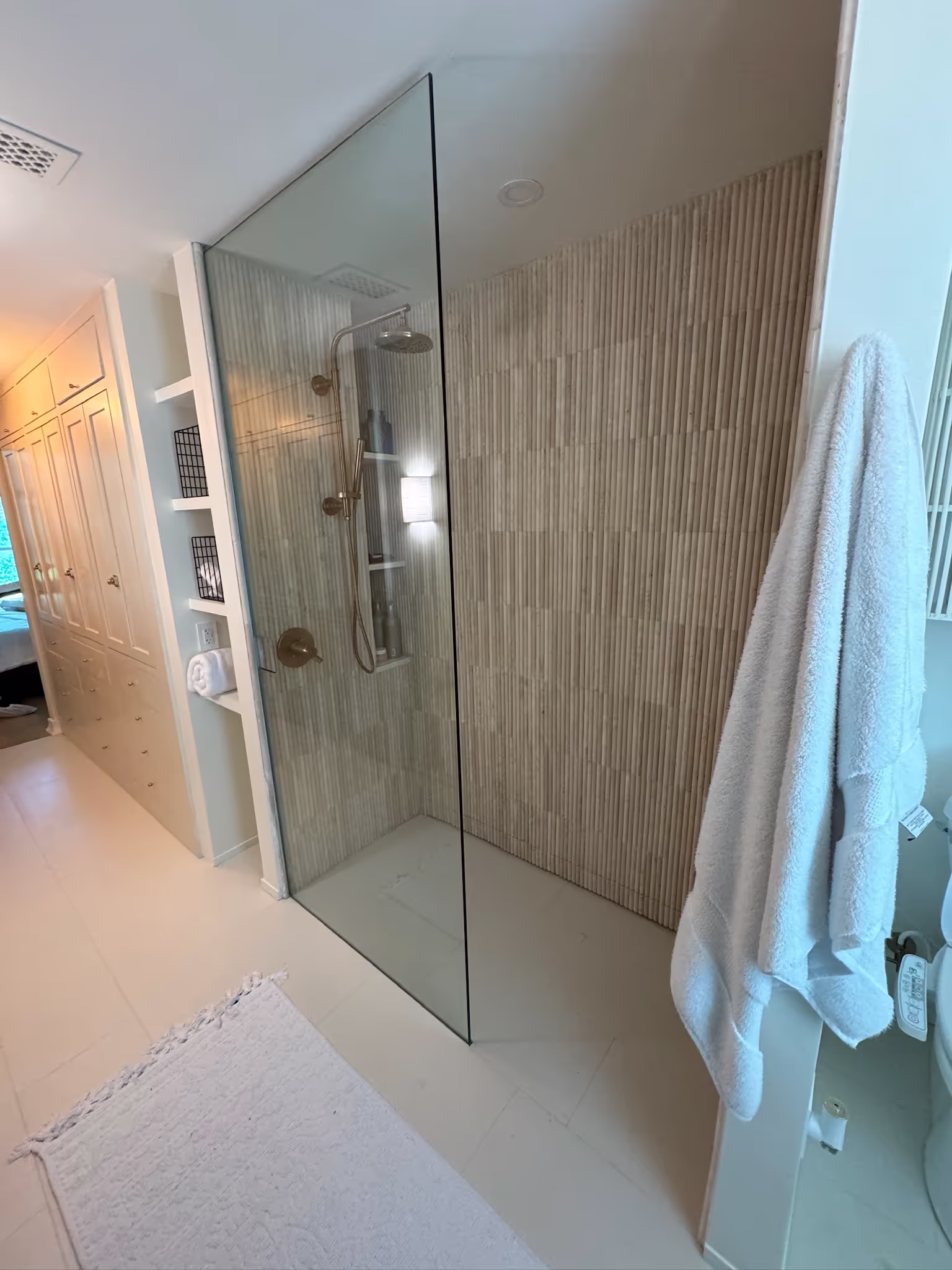 Modern walk-in shower with clear glass panel, vertical textured beige tiles, and chrome shower fixtures in a bright bathroom.