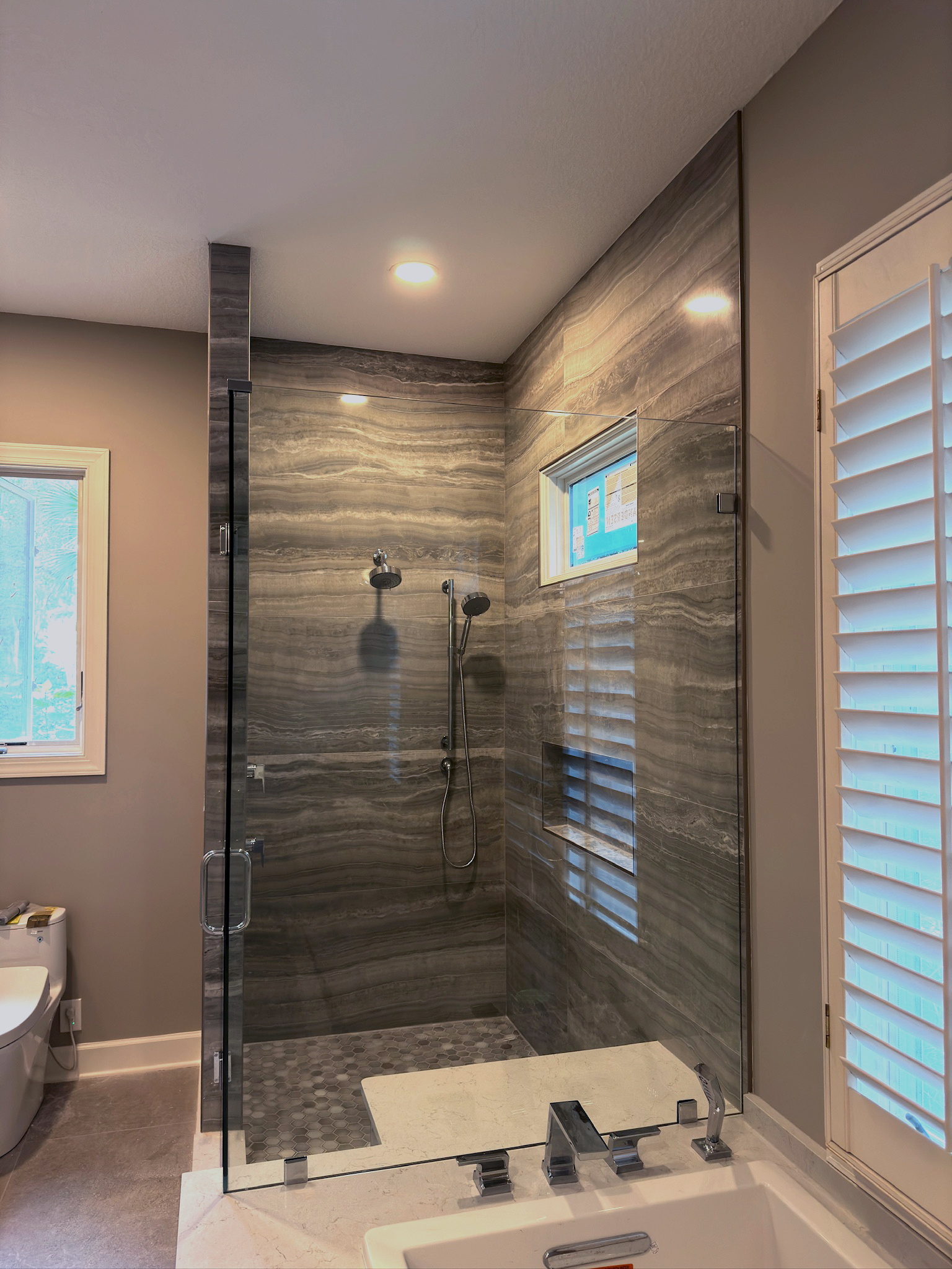 Modern bathroom with glass-enclosed shower featuring gray marble walls, hexagonal floor tiles, and a window with white shutters.
